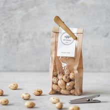 Lade das Bild in den Galerie-Viewer, Ein Beutel Mandeln mit Schokoüberzug von Nicolas Vahé, der mit einer goldenen Klammer verschlossen ist. Im Vordergrund liegen ein paar lose Schoko-Mandeln.