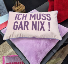 Lade das Bild in den Galerie-Viewer, Ein Kissen mit lilafarbenem "Ich muss gar nix"-Schriftzug liegt auf einem fliederfarbenen Fellkissen.