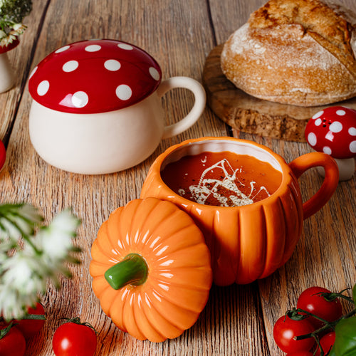 Orange Suppentasse in Kürbisform, gefüllt mit einer Kürbissuppe, der passende Deckel ist an die Schale angelehnt. Um die Tasse herum liegen Tomaten, ein Brot und Fliegenpilz-Deko.