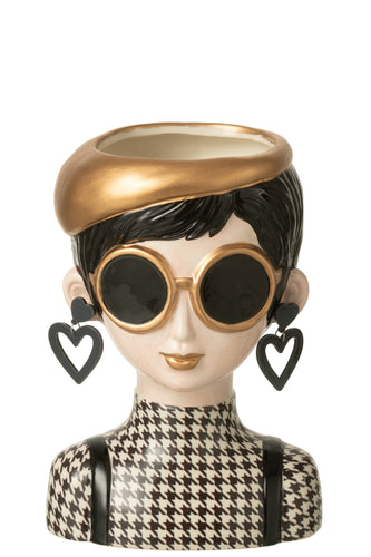 Vase in Form eines Frauenkopfs. Die Frau hat kurze, schwarze Haare, ein schwarz-weißes Oberteil mit Hahnentritt-Muster und trägt dazu eine große Sonnenbrille, Ohrringe  und eine goldene Baskenmütze.