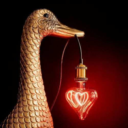 Eine rote Herz-Glühbirne in einer Lampenfassung, deren Kabel im Schnabel einer goldenen Ente hängt.