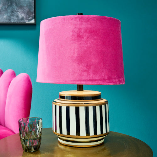 Werner Voss Tischleuchte mit Keramikfuß in schwarz-weiß gestreift und pinkfarbenem Lampenschirm mt Samtbezug. Die Lampe steht auf einem Beistelltisch, daneben ein Glas. IM Hintergrund ein pinker Sessel und eine türkise Wand.