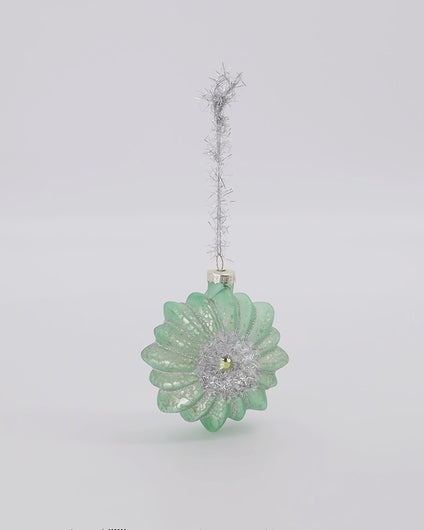 House Doctor Weihnachtsbaumschmuck "Figure aus Glas in Form einer Blüte. Der Anhänger hat eine staubig-grüne Farbe mit silbernen Akzenten, in der Mitte ist silberner Glitter und ein Strassstein.