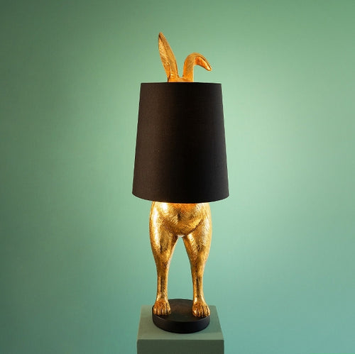 Die Lampe Hiding Bunny von Werner Voss ist ein goldfarbener Hase, der sich hinter einem schwarzen Lampenschirm versteckt. Die Beine und Ohren des Osterhasen gucken hervor. Ideal als Stehlampe im Wohnzimmer oder Leuchte für Ostern und die Osterdeko.
