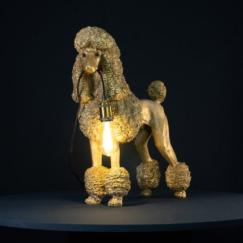 Lampe in Form eines goldenen Pudels von werner Voß. Der Hund hält ein Kabel in der Schnauze an dem eine Fassung für eine Glühbirne hängt.