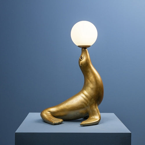 Deko-Lampe Robbie von Werner Voß in Form einer goldenen Seerobbe, die einen Ball - der runde Lampenschirm aus Glas - auf der Schnauze balanciert.